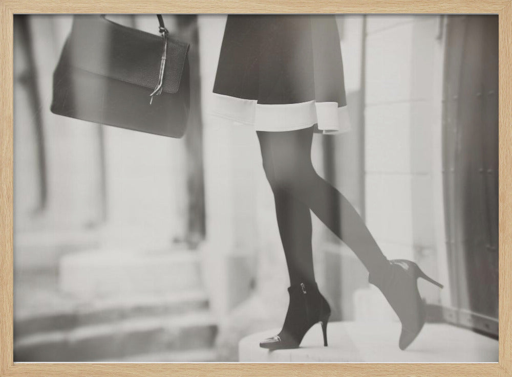 A framed, black and white fashion photograph focusing on the lower half of a woman as she walks. She is wearing a dark skirt with a white hem, black tights, and stylish high-heeled ankle boots, and she is carrying a dark handbag. Print