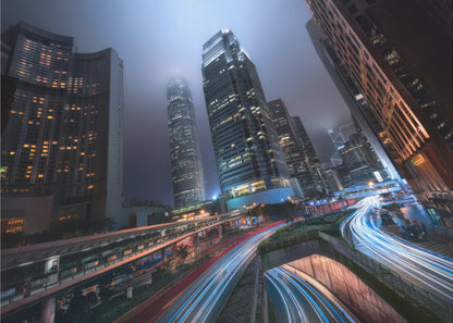 A dynamic, low-angle, long-exposure shot of a modern city at night. Towering skyscrapers with illuminated windows pierce through a foggy, dark blue sky. Below, curving highways are filled with the vibrant red and white light trails of moving traffic. Print