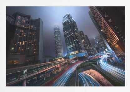 A dynamic, low-angle, long-exposure shot of a modern city at night. Towering skyscrapers with illuminated windows pierce through a foggy, dark blue sky. Below, curving highways are filled with the vibrant red and white light trails of moving traffic. Print