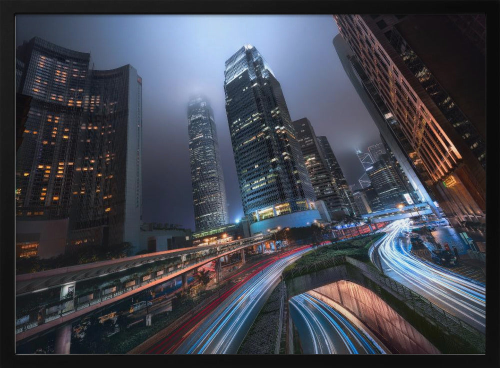A dynamic, low-angle, long-exposure shot of a modern city at night. Towering skyscrapers with illuminated windows pierce through a foggy, dark blue sky. Below, curving highways are filled with the vibrant red and white light trails of moving traffic. Print