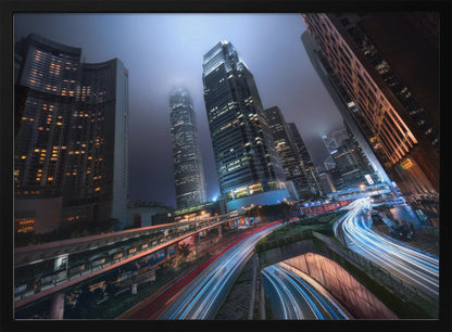A dynamic, low-angle, long-exposure shot of a modern city at night. Towering skyscrapers with illuminated windows pierce through a foggy, dark blue sky. Below, curving highways are filled with the vibrant red and white light trails of moving traffic. Print