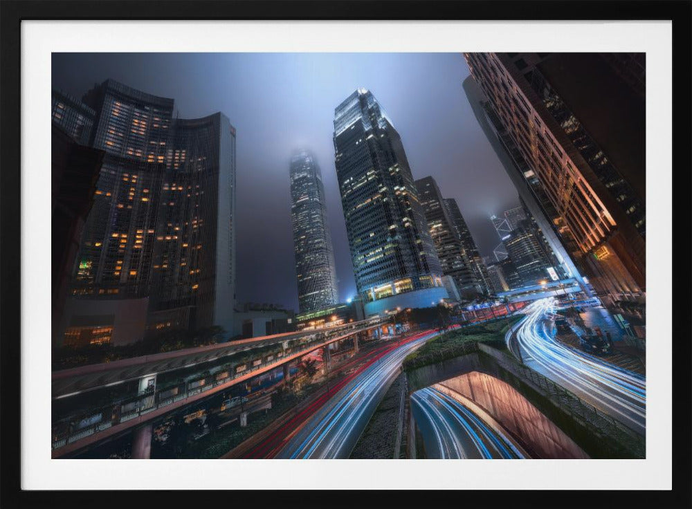 A dynamic, low-angle, long-exposure shot of a modern city at night. Towering skyscrapers with illuminated windows pierce through a foggy, dark blue sky. Below, curving highways are filled with the vibrant red and white light trails of moving traffic. Print