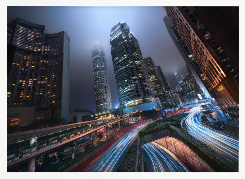 A dynamic, low-angle, long-exposure shot of a modern city at night. Towering skyscrapers with illuminated windows pierce through a foggy, dark blue sky. Below, curving highways are filled with the vibrant red and white light trails of moving traffic. Print