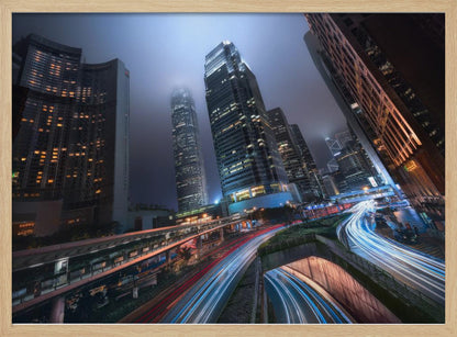 A dynamic, low-angle, long-exposure shot of a modern city at night. Towering skyscrapers with illuminated windows pierce through a foggy, dark blue sky. Below, curving highways are filled with the vibrant red and white light trails of moving traffic. Print