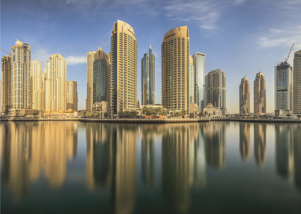 A panoramic photograph of the Dubai Marina skyline with its golden skyscrapers reflecting in the calm, dark water below, all under a clear blue sky and framed in silver. Wall Art