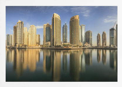 A panoramic photograph of the Dubai Marina skyline with its golden skyscrapers reflecting in the calm, dark water below, all under a clear blue sky and framed in silver. Wall Art