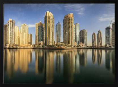 A panoramic photograph of the Dubai Marina skyline with its golden skyscrapers reflecting in the calm, dark water below, all under a clear blue sky and framed in silver. Wall Art