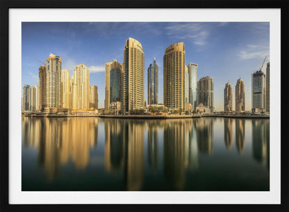 A panoramic photograph of the Dubai Marina skyline with its golden skyscrapers reflecting in the calm, dark water below, all under a clear blue sky and framed in silver. Wall Art