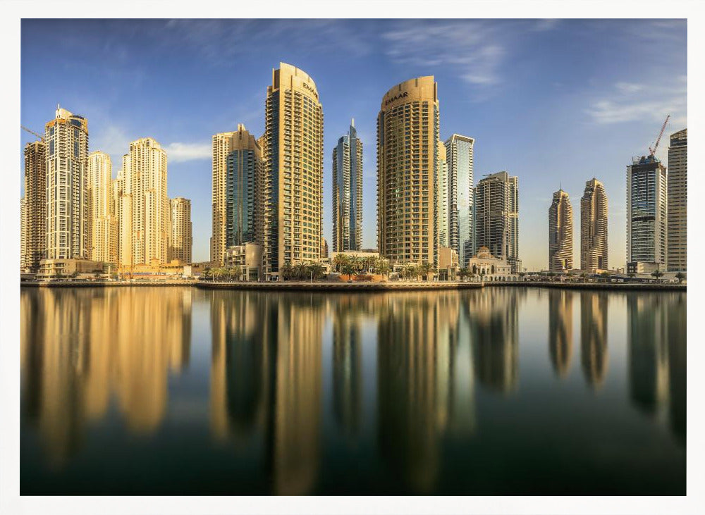 A panoramic photograph of the Dubai Marina skyline with its golden skyscrapers reflecting in the calm, dark water below, all under a clear blue sky and framed in silver. Wall Art