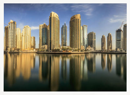 A panoramic photograph of the Dubai Marina skyline with its golden skyscrapers reflecting in the calm, dark water below, all under a clear blue sky and framed in silver. Wall Art