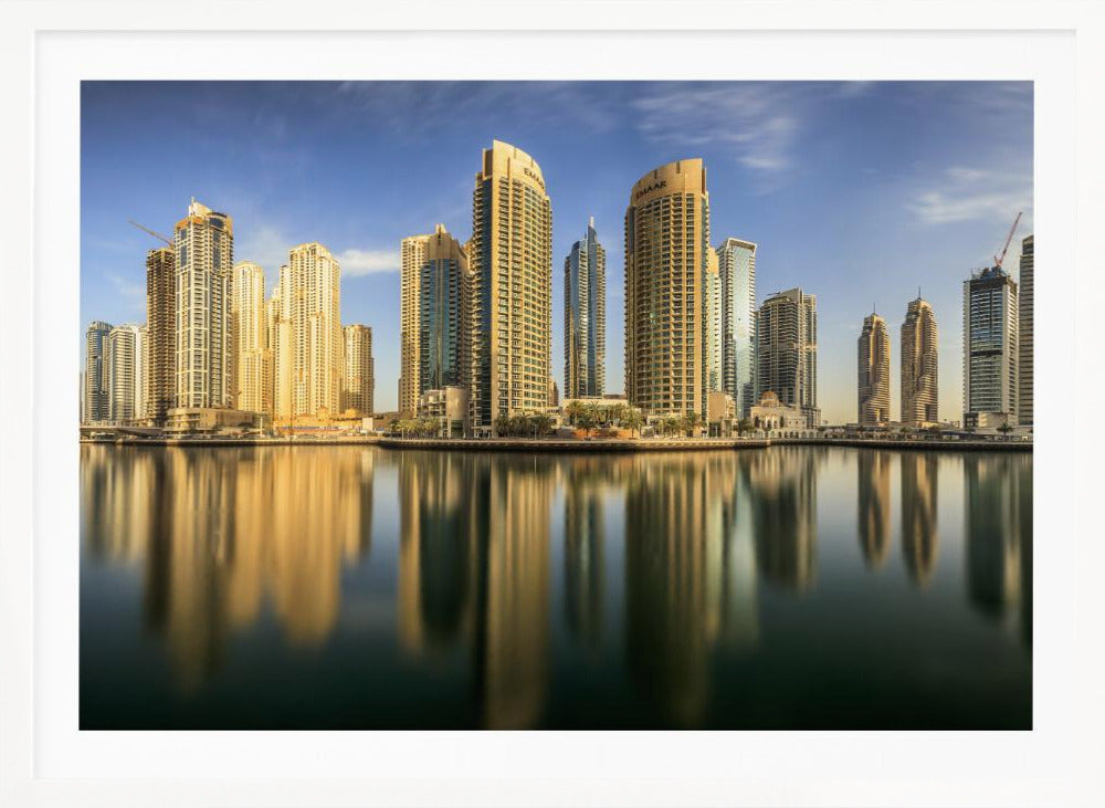 A panoramic photograph of the Dubai Marina skyline with its golden skyscrapers reflecting in the calm, dark water below, all under a clear blue sky and framed in silver. Wall Art