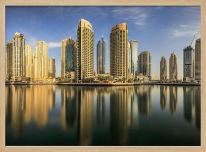 A panoramic photograph of the Dubai Marina skyline with its golden skyscrapers reflecting in the calm, dark water below, all under a clear blue sky and framed in silver. Wall Art