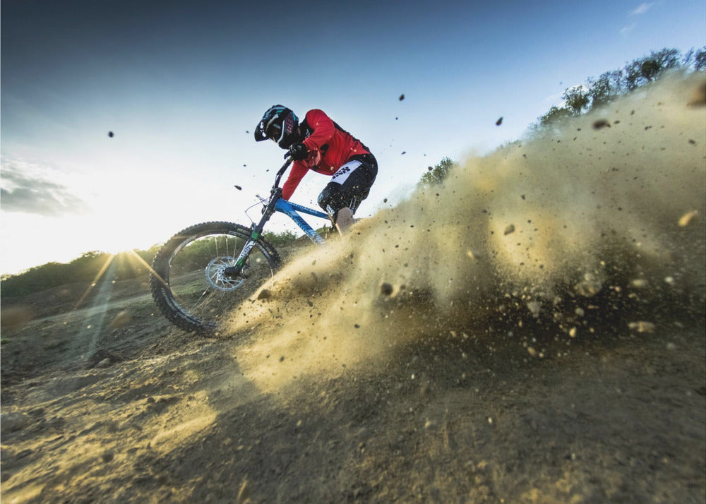 A mountain biker wearing a red jersey and helmet speeds down a steep, dusty trail, kicking up a large cloud of dirt. The sun is setting in the background, creating a lens flare against a clear blue sky. The image is captured from a low angle, emphasizing the action and speed, and is presented within a silver frame. Artwork