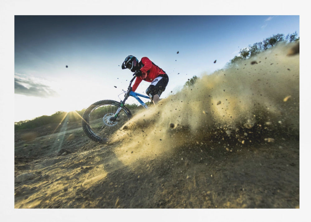 A mountain biker wearing a red jersey and helmet speeds down a steep, dusty trail, kicking up a large cloud of dirt. The sun is setting in the background, creating a lens flare against a clear blue sky. The image is captured from a low angle, emphasizing the action and speed, and is presented within a silver frame. Artwork
