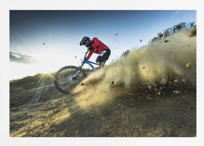 A mountain biker wearing a red jersey and helmet speeds down a steep, dusty trail, kicking up a large cloud of dirt. The sun is setting in the background, creating a lens flare against a clear blue sky. The image is captured from a low angle, emphasizing the action and speed, and is presented within a silver frame. Artwork