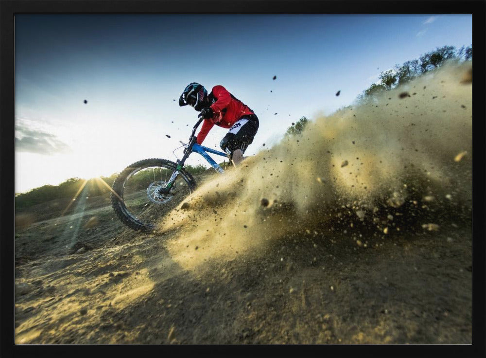A mountain biker wearing a red jersey and helmet speeds down a steep, dusty trail, kicking up a large cloud of dirt. The sun is setting in the background, creating a lens flare against a clear blue sky. The image is captured from a low angle, emphasizing the action and speed, and is presented within a silver frame. Artwork