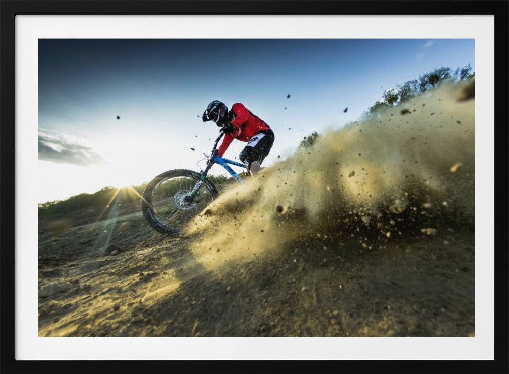 A mountain biker wearing a red jersey and helmet speeds down a steep, dusty trail, kicking up a large cloud of dirt. The sun is setting in the background, creating a lens flare against a clear blue sky. The image is captured from a low angle, emphasizing the action and speed, and is presented within a silver frame. Artwork