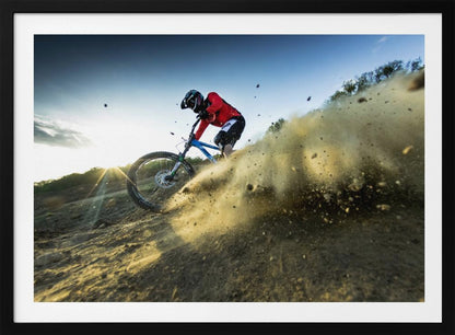 A mountain biker wearing a red jersey and helmet speeds down a steep, dusty trail, kicking up a large cloud of dirt. The sun is setting in the background, creating a lens flare against a clear blue sky. The image is captured from a low angle, emphasizing the action and speed, and is presented within a silver frame. Artwork