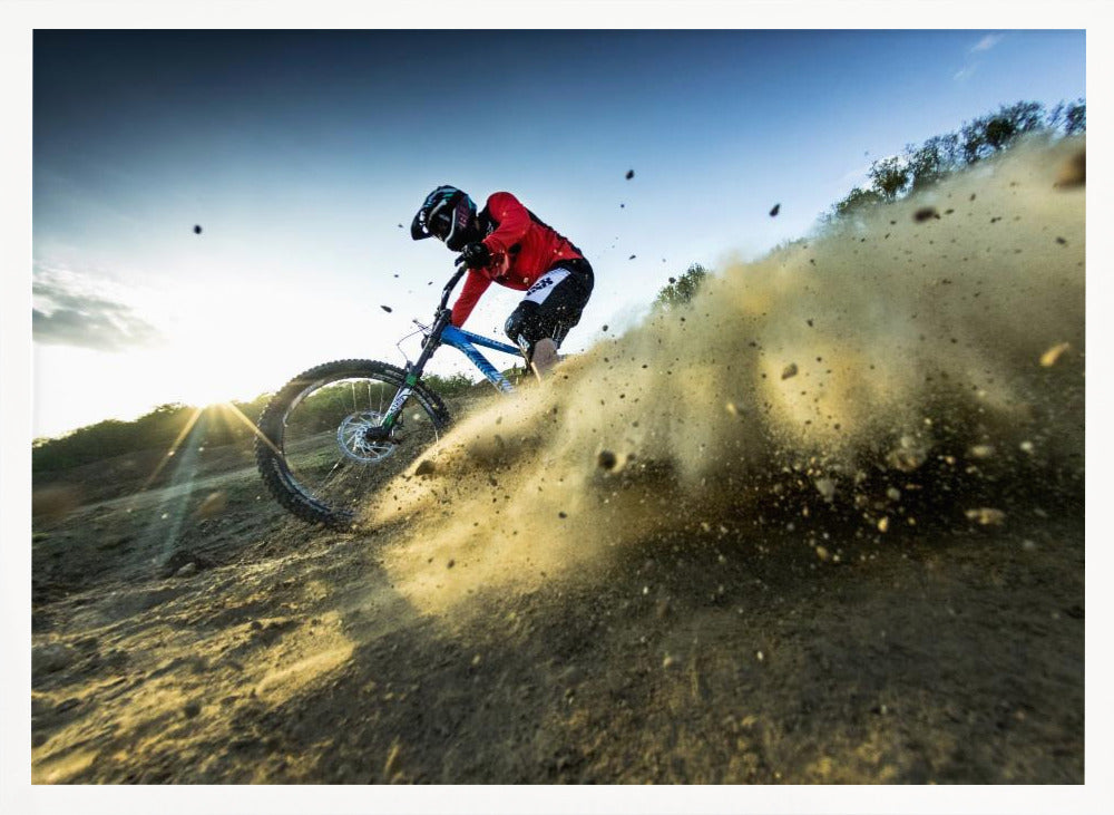 A mountain biker wearing a red jersey and helmet speeds down a steep, dusty trail, kicking up a large cloud of dirt. The sun is setting in the background, creating a lens flare against a clear blue sky. The image is captured from a low angle, emphasizing the action and speed, and is presented within a silver frame. Artwork