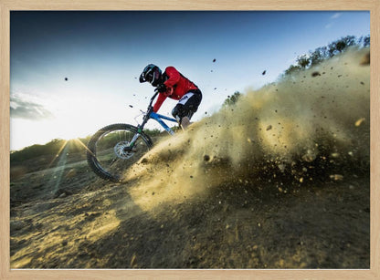 A mountain biker wearing a red jersey and helmet speeds down a steep, dusty trail, kicking up a large cloud of dirt. The sun is setting in the background, creating a lens flare against a clear blue sky. The image is captured from a low angle, emphasizing the action and speed, and is presented within a silver frame. Artwork