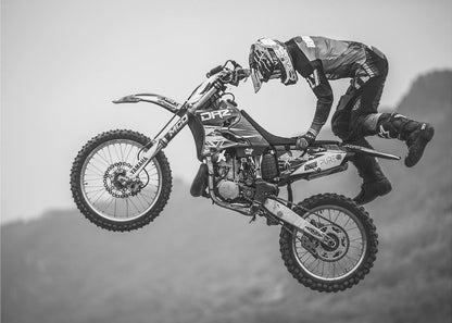 A dynamic black and white photograph of a motocross rider in mid-air, performing a jump. The rider, wearing full protective gear and a helmet, is leaning forward over the handlebars of the dirt bike. The bike is suspended against a blurry, light gray background, emphasizing the action and height of the jump. The image is enclosed in a silver frame. Wall Art