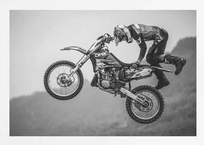 A dynamic black and white photograph of a motocross rider in mid-air, performing a jump. The rider, wearing full protective gear and a helmet, is leaning forward over the handlebars of the dirt bike. The bike is suspended against a blurry, light gray background, emphasizing the action and height of the jump. The image is enclosed in a silver frame. Wall Art