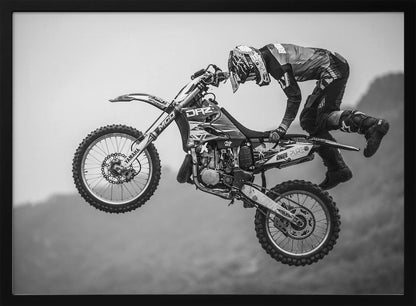 A dynamic black and white photograph of a motocross rider in mid-air, performing a jump. The rider, wearing full protective gear and a helmet, is leaning forward over the handlebars of the dirt bike. The bike is suspended against a blurry, light gray background, emphasizing the action and height of the jump. The image is enclosed in a silver frame. Wall Art