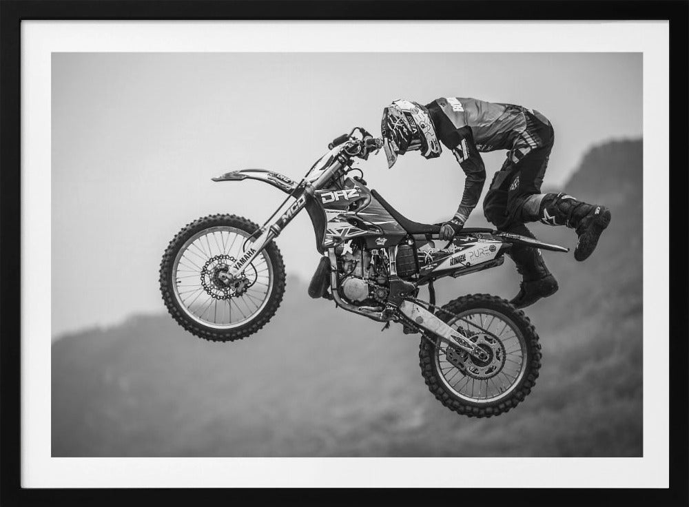 A dynamic black and white photograph of a motocross rider in mid-air, performing a jump. The rider, wearing full protective gear and a helmet, is leaning forward over the handlebars of the dirt bike. The bike is suspended against a blurry, light gray background, emphasizing the action and height of the jump. The image is enclosed in a silver frame. Wall Art