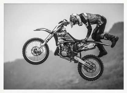 A dynamic black and white photograph of a motocross rider in mid-air, performing a jump. The rider, wearing full protective gear and a helmet, is leaning forward over the handlebars of the dirt bike. The bike is suspended against a blurry, light gray background, emphasizing the action and height of the jump. The image is enclosed in a silver frame. Wall Art