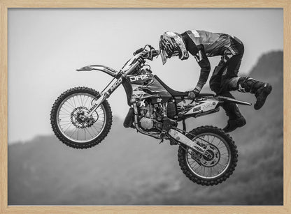 A dynamic black and white photograph of a motocross rider in mid-air, performing a jump. The rider, wearing full protective gear and a helmet, is leaning forward over the handlebars of the dirt bike. The bike is suspended against a blurry, light gray background, emphasizing the action and height of the jump. The image is enclosed in a silver frame. Wall Art