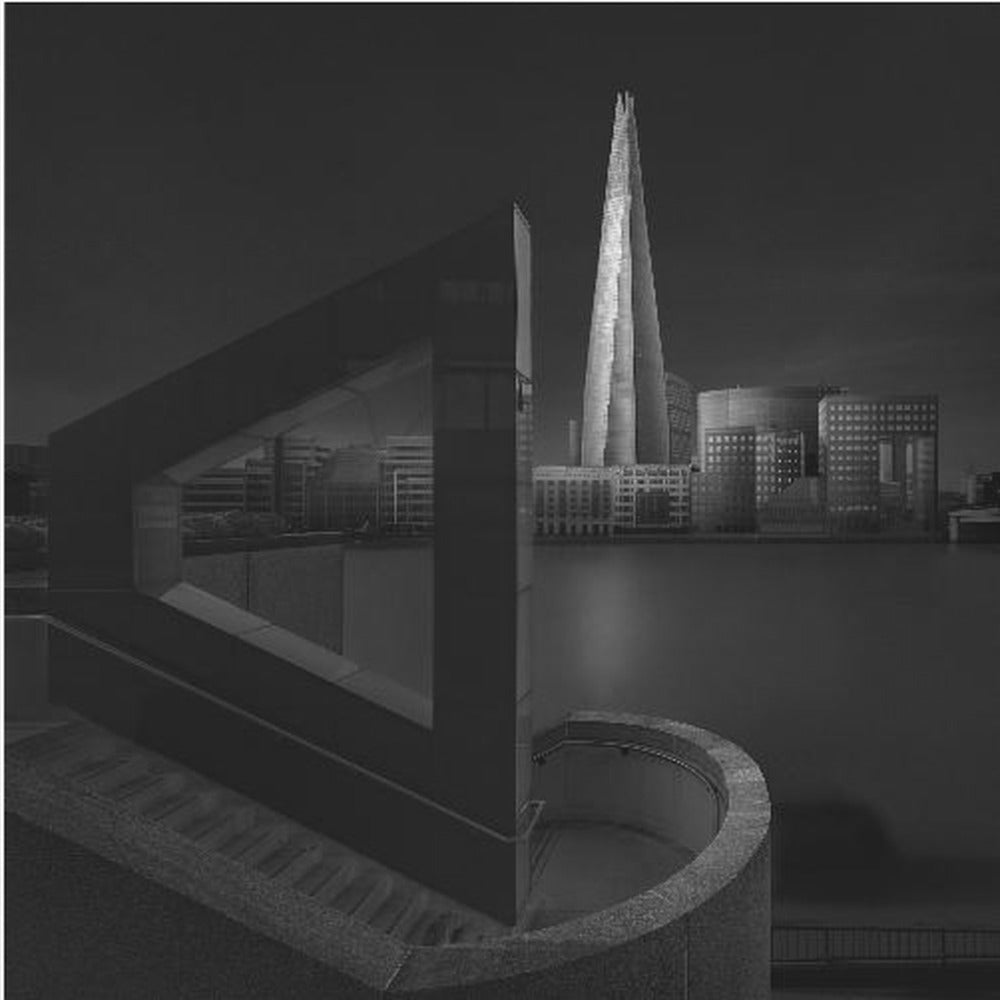 A dramatic black and white architectural photograph of the London skyline at night. The illuminated Shard skyscraper stands tall in the background across a dark river. The foreground features strong geometric shapes, including a large triangular structure and a curved concrete staircase, creating a layered and modern composition. Print