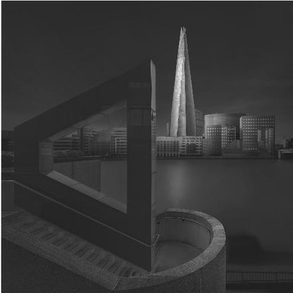 A dramatic black and white architectural photograph of the London skyline at night. The illuminated Shard skyscraper stands tall in the background across a dark river. The foreground features strong geometric shapes, including a large triangular structure and a curved concrete staircase, creating a layered and modern composition. Print
