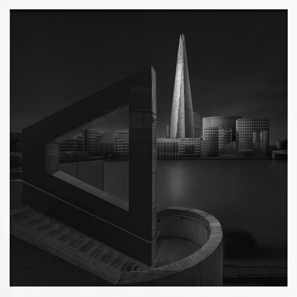 A dramatic black and white architectural photograph of the London skyline at night. The illuminated Shard skyscraper stands tall in the background across a dark river. The foreground features strong geometric shapes, including a large triangular structure and a curved concrete staircase, creating a layered and modern composition. Print