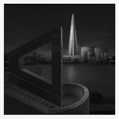 A dramatic black and white architectural photograph of the London skyline at night. The illuminated Shard skyscraper stands tall in the background across a dark river. The foreground features strong geometric shapes, including a large triangular structure and a curved concrete staircase, creating a layered and modern composition. Print