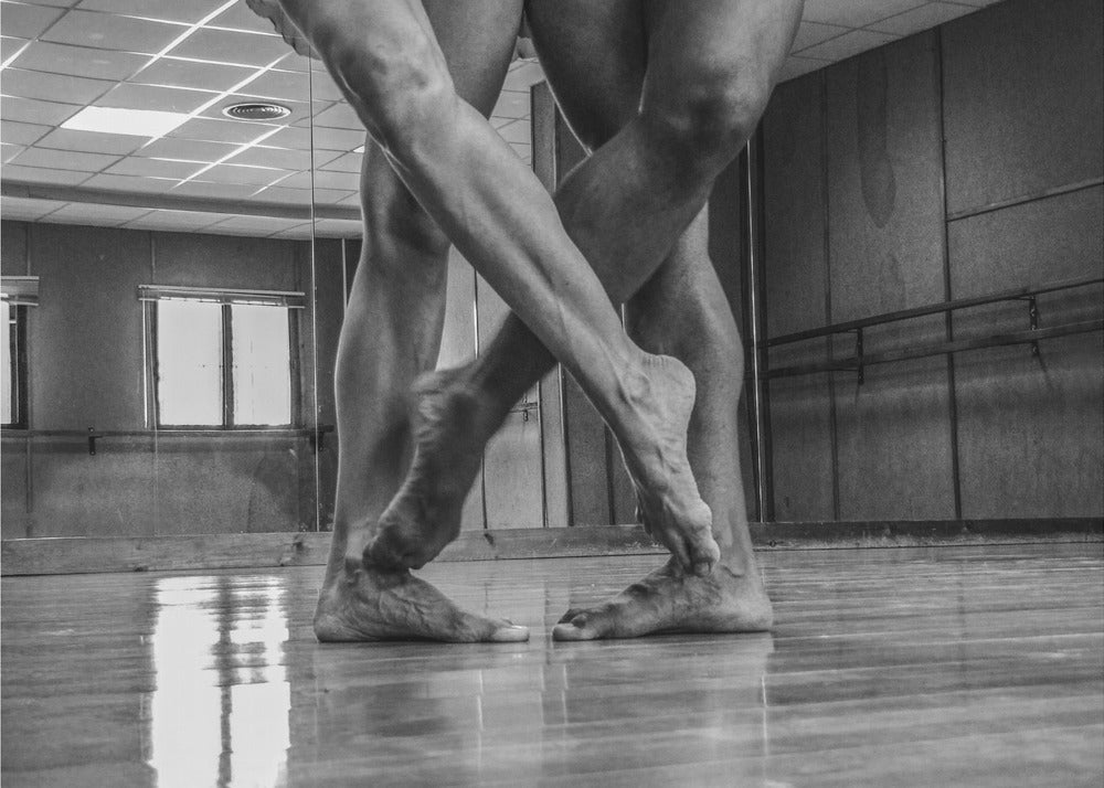 A dramatic low-angle, black and white photograph capturing the muscular legs and bare feet of two dancers intertwined in a dance studio. Their limbs cross in a display of strength and grace on a reflective wooden floor, with the studio's mirrored wall and barre visible in the background. The image is enclosed in a silver-grey frame. Decor