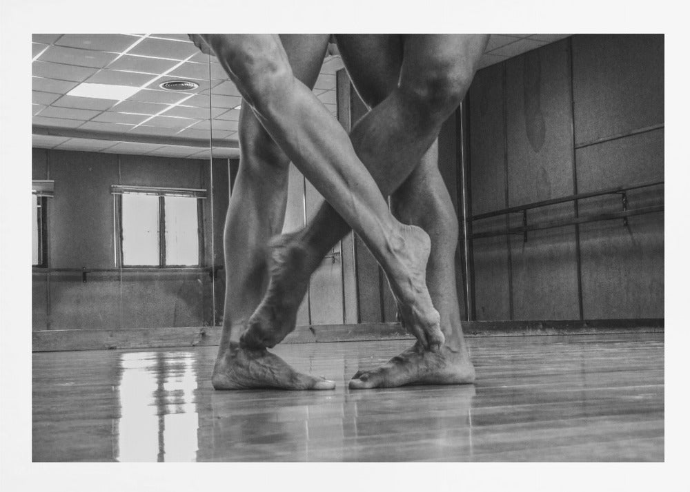 A dramatic low-angle, black and white photograph capturing the muscular legs and bare feet of two dancers intertwined in a dance studio. Their limbs cross in a display of strength and grace on a reflective wooden floor, with the studio's mirrored wall and barre visible in the background. The image is enclosed in a silver-grey frame. Decor