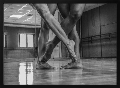 A dramatic low-angle, black and white photograph capturing the muscular legs and bare feet of two dancers intertwined in a dance studio. Their limbs cross in a display of strength and grace on a reflective wooden floor, with the studio's mirrored wall and barre visible in the background. The image is enclosed in a silver-grey frame. Decor
