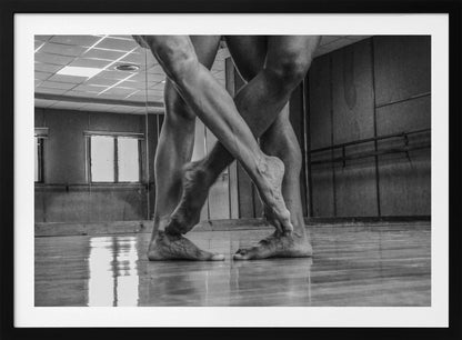 A dramatic low-angle, black and white photograph capturing the muscular legs and bare feet of two dancers intertwined in a dance studio. Their limbs cross in a display of strength and grace on a reflective wooden floor, with the studio's mirrored wall and barre visible in the background. The image is enclosed in a silver-grey frame. Decor