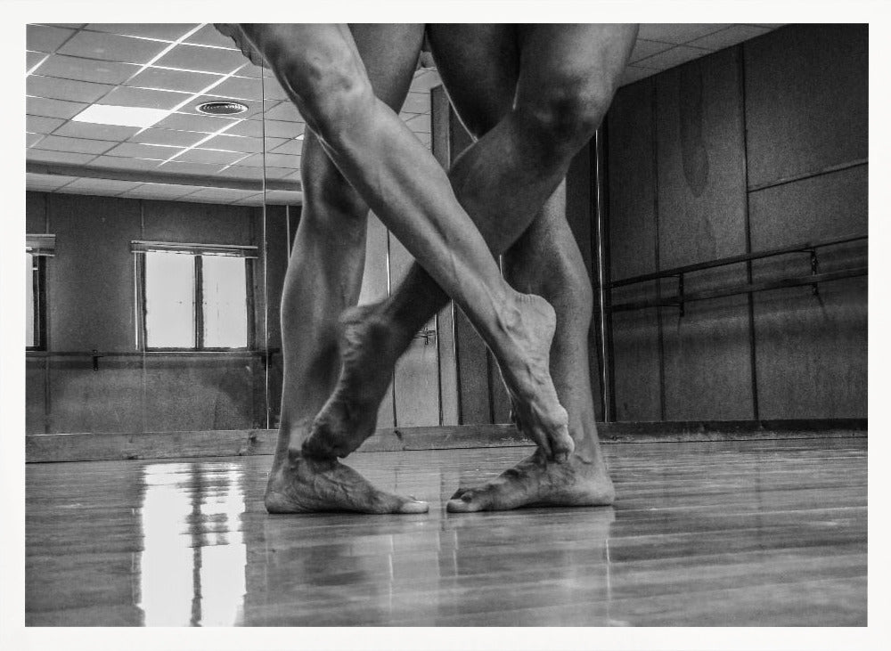 A dramatic low-angle, black and white photograph capturing the muscular legs and bare feet of two dancers intertwined in a dance studio. Their limbs cross in a display of strength and grace on a reflective wooden floor, with the studio's mirrored wall and barre visible in the background. The image is enclosed in a silver-grey frame. Decor