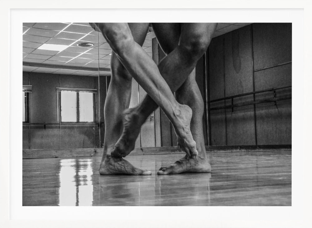 A dramatic low-angle, black and white photograph capturing the muscular legs and bare feet of two dancers intertwined in a dance studio. Their limbs cross in a display of strength and grace on a reflective wooden floor, with the studio's mirrored wall and barre visible in the background. The image is enclosed in a silver-grey frame. Decor
