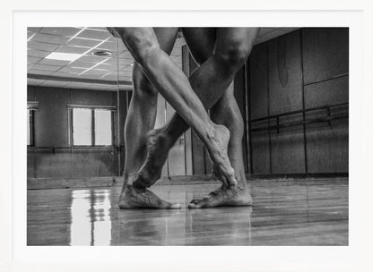 A dramatic low-angle, black and white photograph capturing the muscular legs and bare feet of two dancers intertwined in a dance studio. Their limbs cross in a display of strength and grace on a reflective wooden floor, with the studio's mirrored wall and barre visible in the background. The image is enclosed in a silver-grey frame. Decor