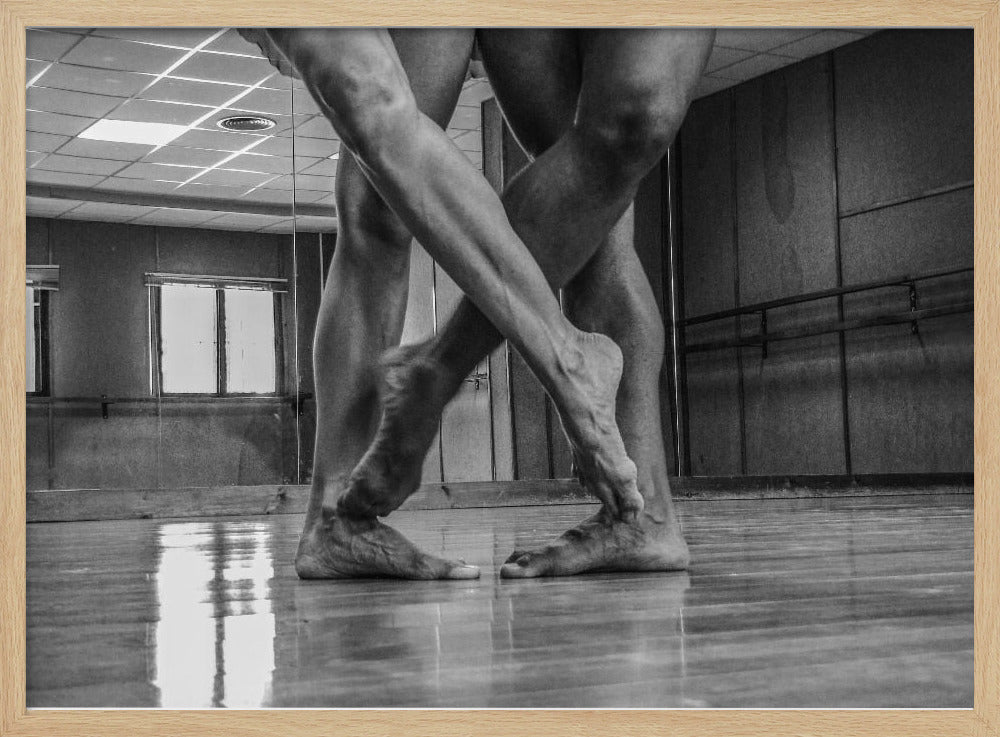 A dramatic low-angle, black and white photograph capturing the muscular legs and bare feet of two dancers intertwined in a dance studio. Their limbs cross in a display of strength and grace on a reflective wooden floor, with the studio's mirrored wall and barre visible in the background. The image is enclosed in a silver-grey frame. Decor