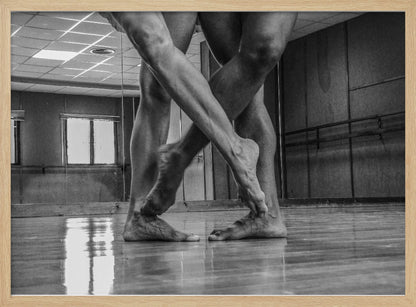 A dramatic low-angle, black and white photograph capturing the muscular legs and bare feet of two dancers intertwined in a dance studio. Their limbs cross in a display of strength and grace on a reflective wooden floor, with the studio's mirrored wall and barre visible in the background. The image is enclosed in a silver-grey frame. Decor