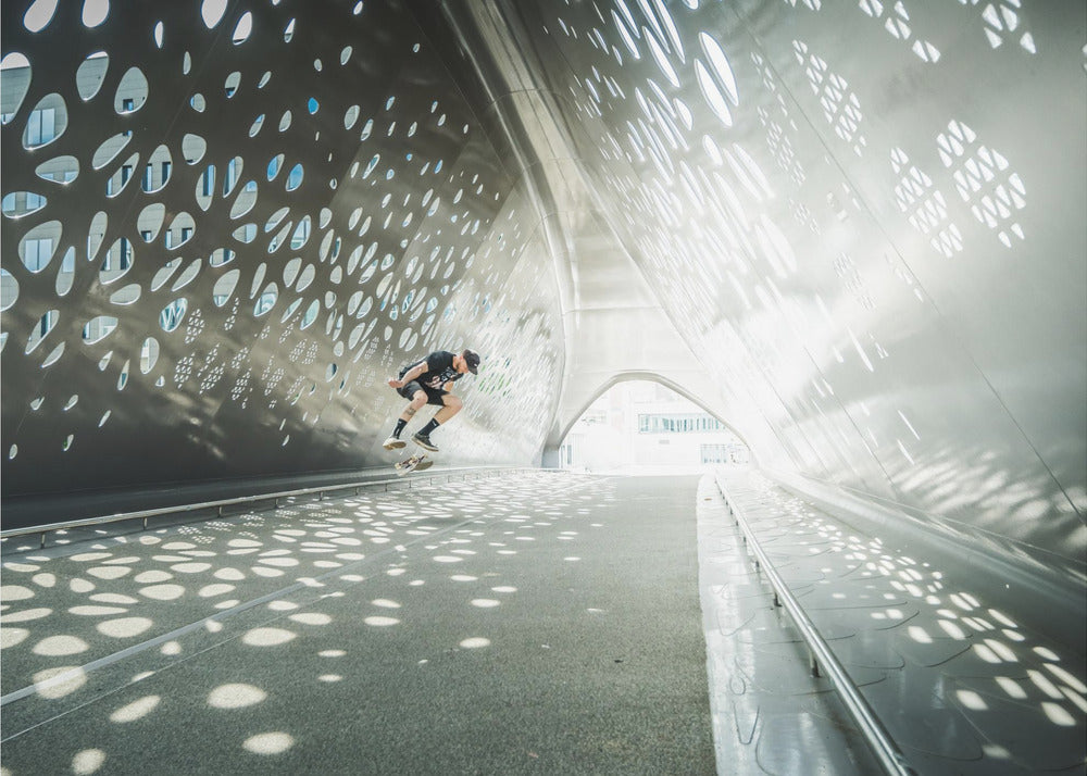 A skateboarder is captured mid-air performing a trick inside a futuristic, metallic tunnel with patterned cutouts that cast dappled light and shadows across the scene, all enclosed within a silver frame. Artwork