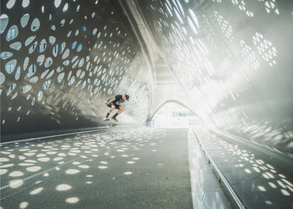 A skateboarder is captured mid-air performing a trick inside a futuristic, metallic tunnel with patterned cutouts that cast dappled light and shadows across the scene, all enclosed within a silver frame. Artwork