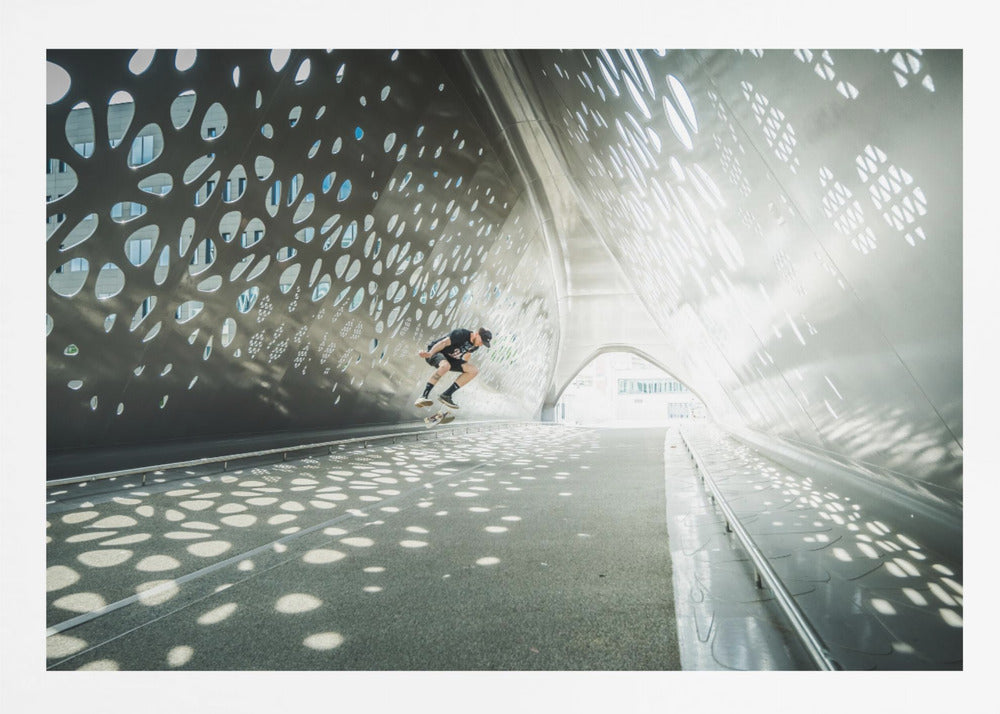 A skateboarder is captured mid-air performing a trick inside a futuristic, metallic tunnel with patterned cutouts that cast dappled light and shadows across the scene, all enclosed within a silver frame. Artwork