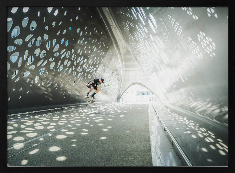 A skateboarder is captured mid-air performing a trick inside a futuristic, metallic tunnel with patterned cutouts that cast dappled light and shadows across the scene, all enclosed within a silver frame. Artwork