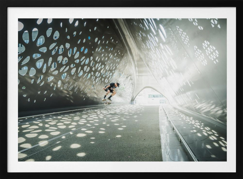 A skateboarder is captured mid-air performing a trick inside a futuristic, metallic tunnel with patterned cutouts that cast dappled light and shadows across the scene, all enclosed within a silver frame. Artwork