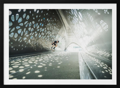 A skateboarder is captured mid-air performing a trick inside a futuristic, metallic tunnel with patterned cutouts that cast dappled light and shadows across the scene, all enclosed within a silver frame. Artwork