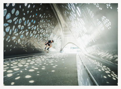 A skateboarder is captured mid-air performing a trick inside a futuristic, metallic tunnel with patterned cutouts that cast dappled light and shadows across the scene, all enclosed within a silver frame. Artwork